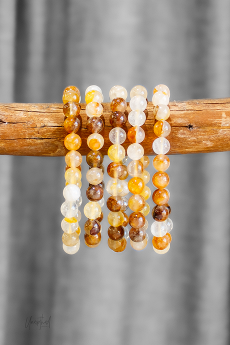 Golden Healer beaded bracelets in yellow, brown, and white on a wooden stick against a gray background.