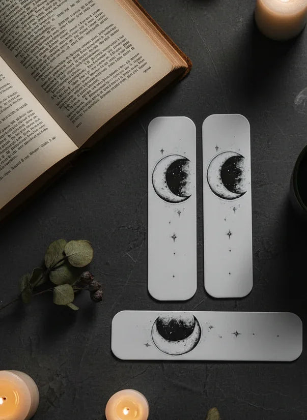 Three PLASTIC bookmarks with moon designs on a dark surface with candles and an open book.