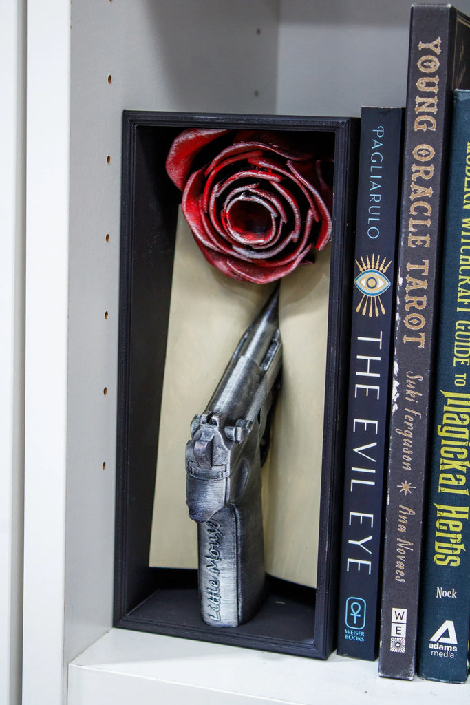 Decorative bookend shaped like a gun and a red rose on a shelf with books.