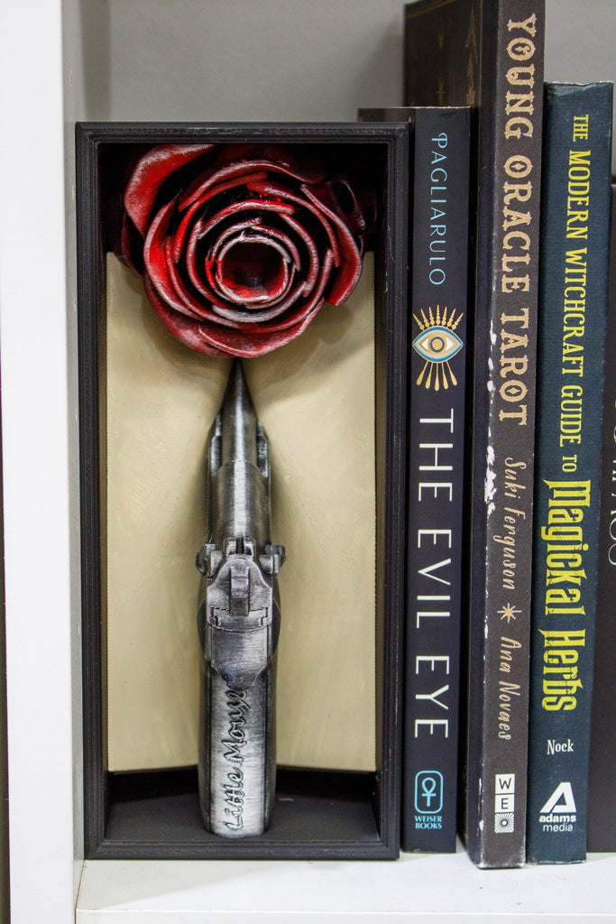 Decorative item with a red rose and silver dagger in a box, placed next to books on a shelf.