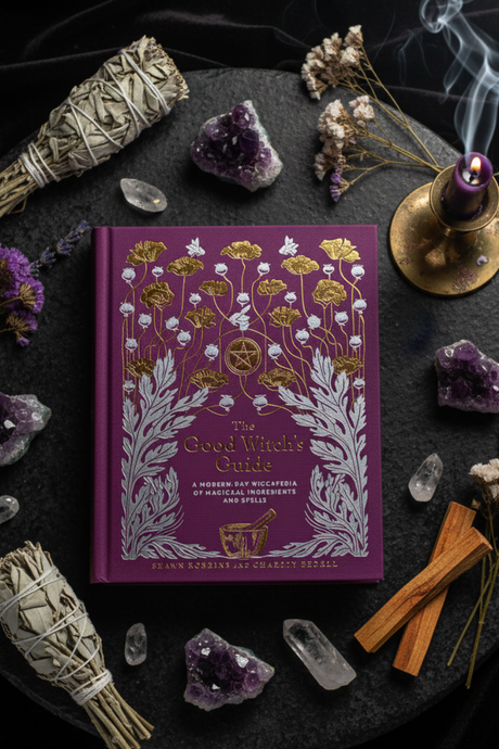 Purple book titled 'The Good Witch's Guide' surrounded by crystals, smudge sticks, and a candle on a dark surface.