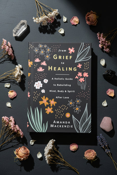 A book titled "from Grief to Healing" by Amanda Mackenzie lies flat on a dark charcoal surface, surrounded by dried flowers, petals, crystals, and small stones. The cover is dark gray with white text and botanical illustrations in pink, orange, and light gray.