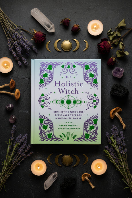 Book titled 'The Holistic Witch' surrounded by candles, crystals, and herbs on a dark background