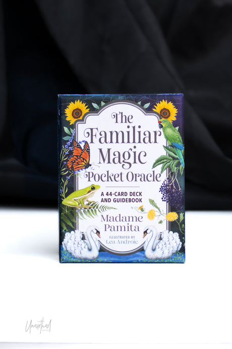 The Familiar Magic Pocket Oracle card deck box by Madame Pamita, featuring a colorful vintage-style illustration with swans, a green parrot, a butterfly, sunflowers, and purple berries, photographed standing upright on a white surface against a dark draped background.