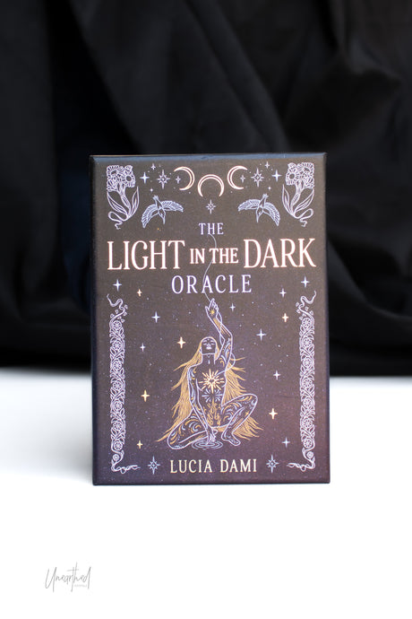 The Light in the Dark Oracle card deck box by Lucia Dami, featuring a dark brown cover with silver celestial illustrations, set against a black draped background on a white surface.