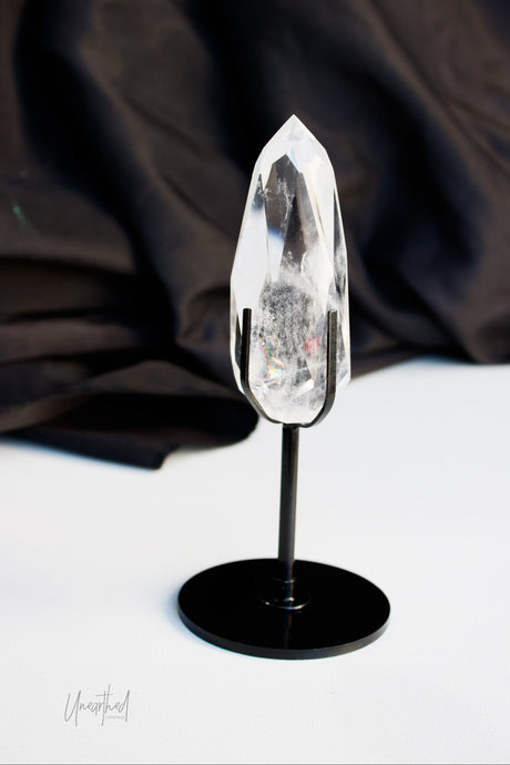 A clear pointed crystal specimen mounted on a black display stand, photographed against a dark draped fabric backdrop and white surface.