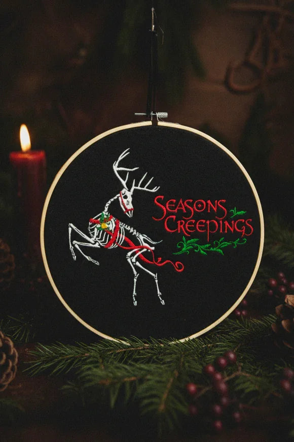 Embroidery hoop with a skeleton deer design and 'Seasons Creepings' text, surrounded by Christmas decorations.