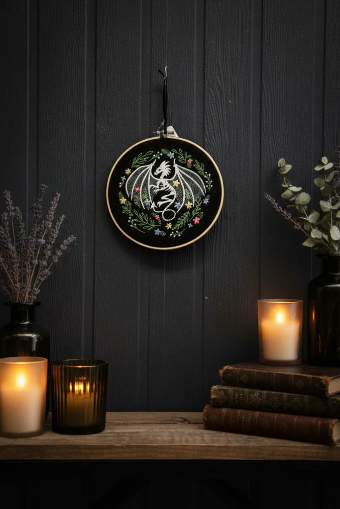 Decorative embroidery hoop with a floral design on a dark wooden wall with candles and books.