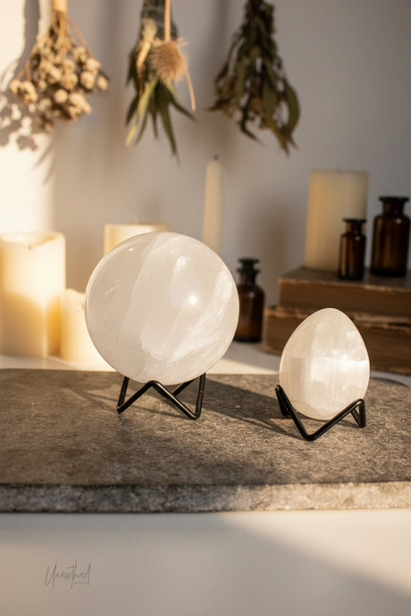 Two white polished crystal spheres on black metal stands, displayed on a dark gray slate board against a draped white fabric background.