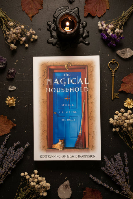 The book cover of 'The Magical Household' by Scott Cunningham & David Harrington, featuring a vibrant blue door illustration with a cat and broom on the cover, displayed on a gray stone surface against a black fabric backdrop.