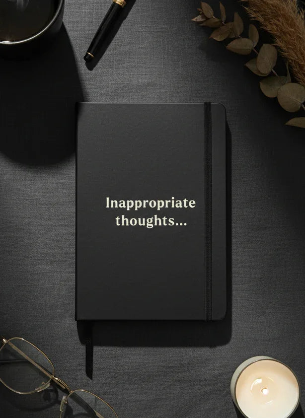 Black notebook with 'inappropriate thoughts...' text on a dark surface with glasses, candle, and leaves.