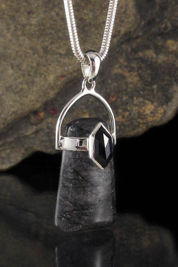 A silver pendant necklace featuring a polished black stone with a faceted leaf-like cut, suspended from a silver chain. The pendant is displayed against a dark, textured rock and a reflective black surface.