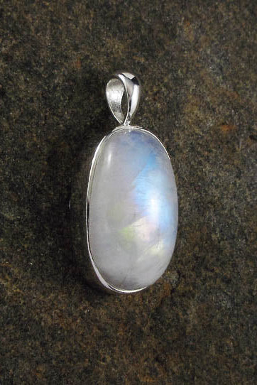 A silver pendant with an oval-shaped iridescent moonstone gemstone, displayed on a dark textured stone surface.