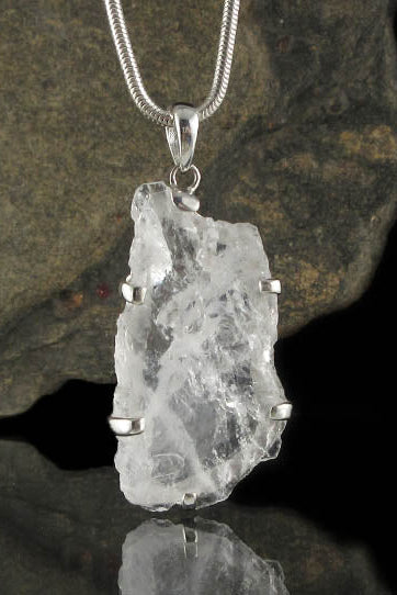 A raw, rough-cut clear quartz crystal pendant held in silver prongs on a silver snake chain, displayed against a dark textured rock with a reflective black surface below.