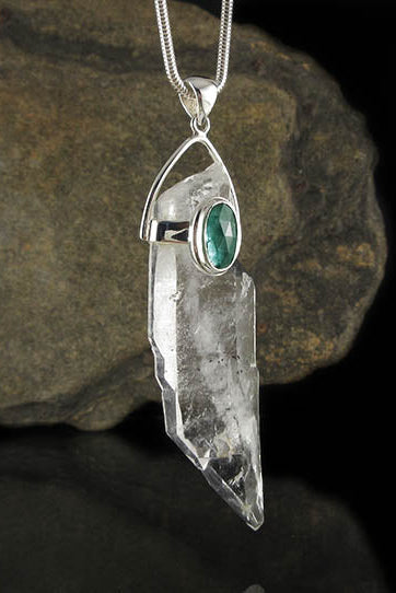 A silver pendant necklace featuring a clear crystal point with a small oval green stone accent, hanging against a dark rock and black background.