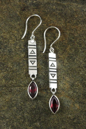 A pair of sterling silver dangle earrings with geometric engraved details and marquise-cut garnet gemstones, displayed on a textured dark brown stone surface.