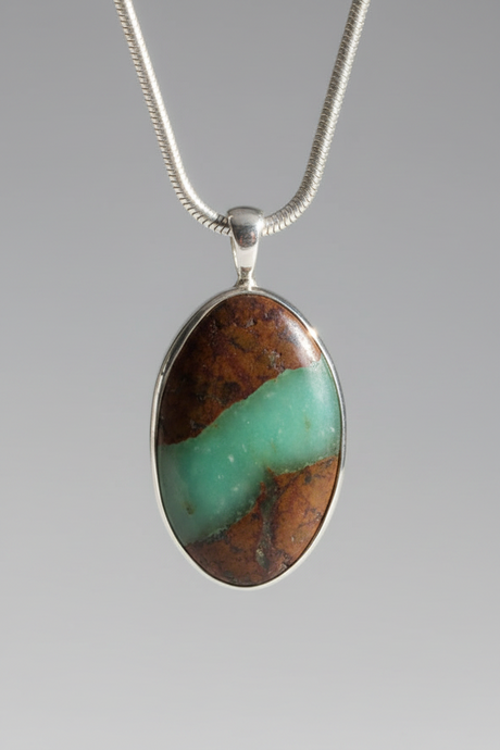 An oval pendant necklace featuring a natural brown and turquoise stone in a silver bezel setting on a silver snake chain, shown against a neutral gray gradient background.