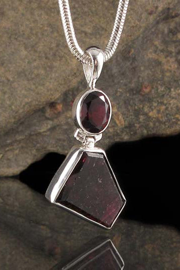 A silver pendant necklace featuring a large faceted dark red gemstone and a smaller oval dark red gemstone, displayed against a dark textured rock background.