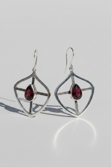 A pair of silver geometric dangle earrings with teardrop-shaped garnet gemstones, displayed on a light gray reflective surface.