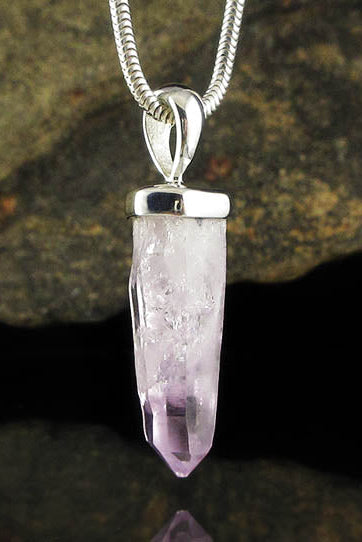 A silver pendant necklace featuring a faceted light purple crystal point hanging from a silver snake chain, set against a dark reflective surface with a blurred brown rock background.