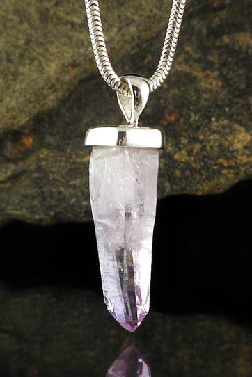 A silver chain necklace with a raw amethyst crystal point pendant, shown hanging against a dark, reflective rock surface.