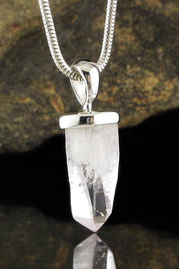 A silver-toned snake chain necklace with a clear faceted crystal point pendant, shown against a dark reflective surface with a blurred rock in the background.