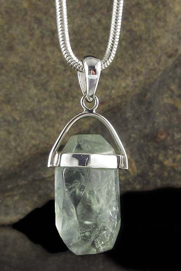 A silver pendant necklace featuring a faceted, point-cut clear quartz crystal held in a silver dome setting, suspended from a silver snake chain against a dark, textured rock background.