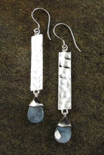 A pair of handmade sterling silver dangle earrings with hammered rectangular bars and teardrop-shaped blue stones, displayed on a dark textured stone background.