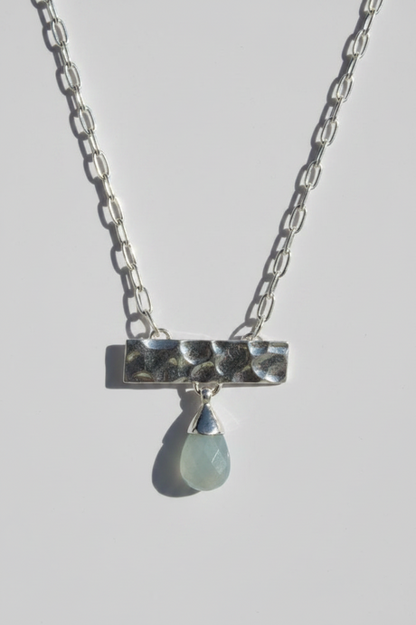 A silver chain necklace with a hammered rectangular bar pendant and a dangling teardrop-shaped pale green stone, shown on a light gray background.