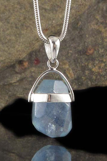 A silver pendant necklace featuring a faceted light blue gemstone in a silver dome setting, suspended from a silver snake chain against a dark reflective background.