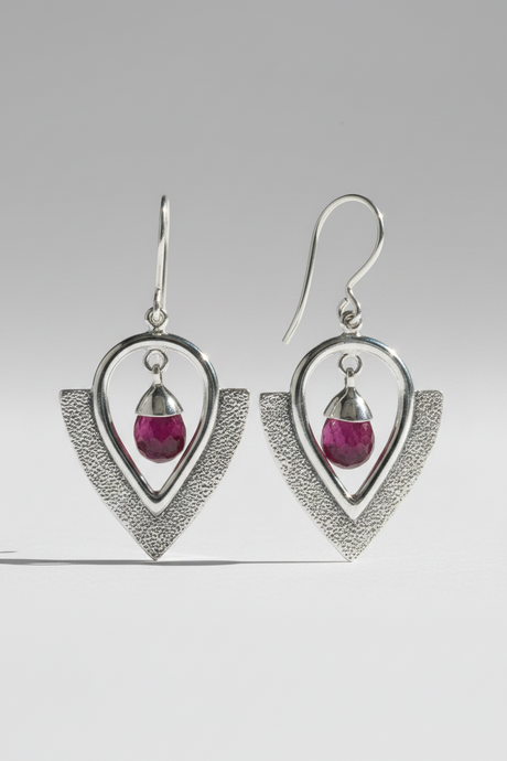 A pair of silver dangle earrings with textured and smooth geometric heart-shaped frames, each holding a small faceted magenta-pink teardrop gemstone, shown on a neutral gray background.
