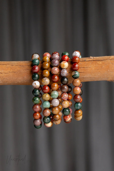 Multicolored Ocean Jasper beaded bracelet on a wooden stick against a gray background