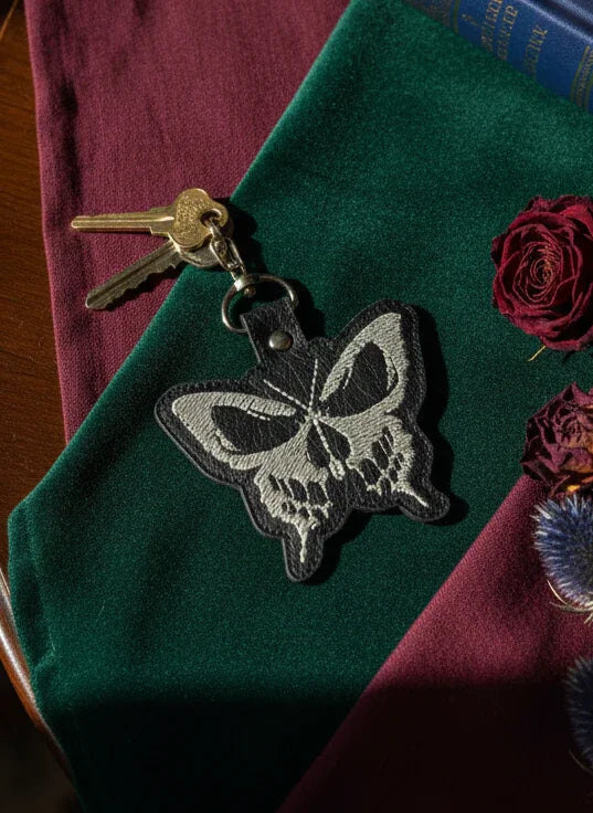 Butterfly-shaped keychain with skull design on a green surface with flowers and keys.