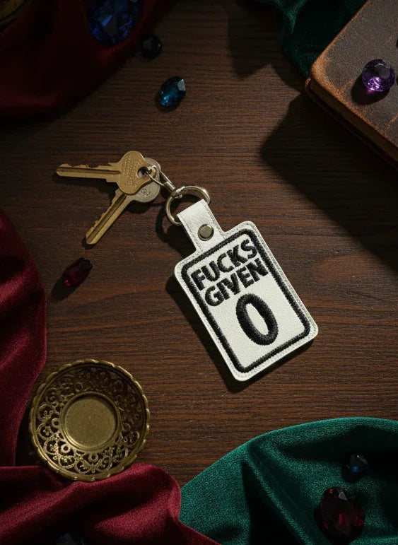 Keychain with 'Fucks Given 0' text on a wooden surface with keys and jewelry.