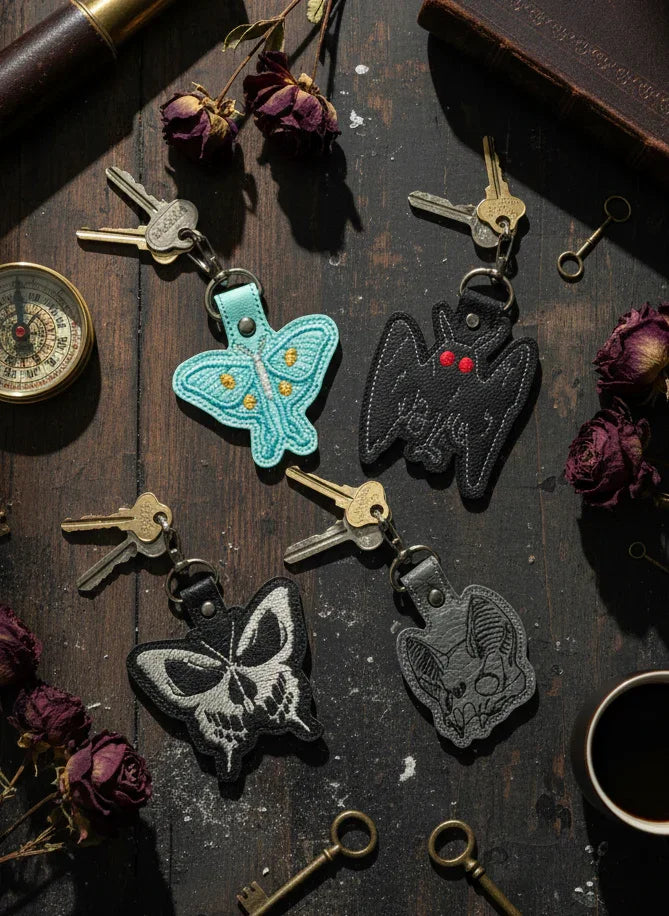 Decorative keychains with butterfly designs on a wooden surface with keys and a cup.