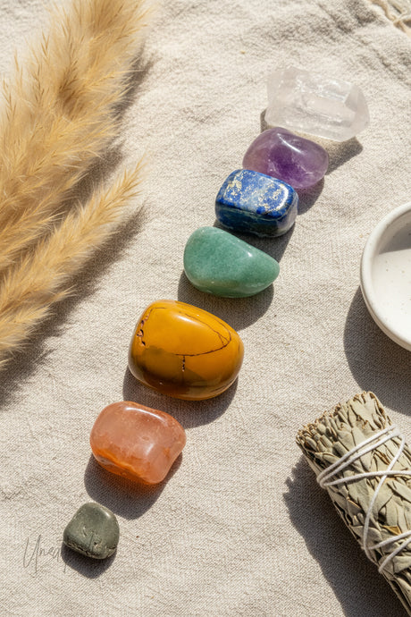 Set of colorful tumbled chakra stones on a textured surface with a bundle of sage and dried grasses.