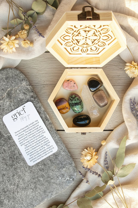 Hexagonal wooden box with stones to assist with grief and a card on a decorative surface with flowers and leaves.