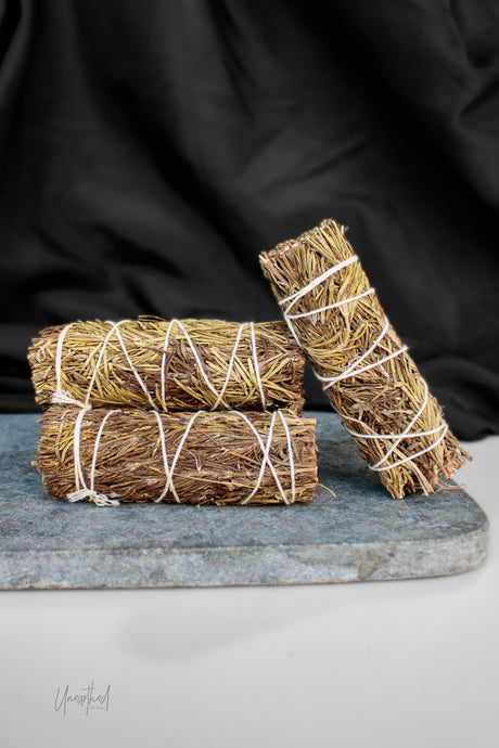 Three bundles of dried rosemary or sage smudge sticks tied with white string, arranged on a gray stone slab against a black fabric backdrop.