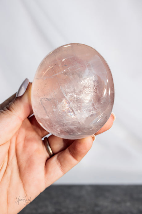 A hand holding a polished, oval-shaped clear crystal sphere with internal inclusions, set against a neutral light gray background.