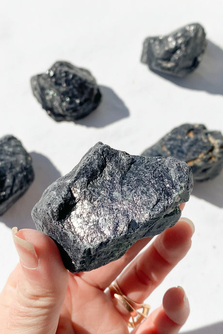 Hand holding a rough Black Tourmaline stone with other stones in the background on a light surface