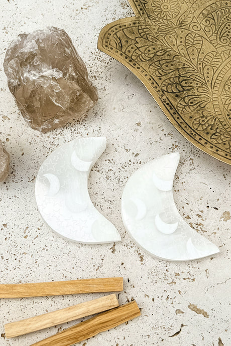 Two white selenite moon-shaped objects on a textured surface with a crystal and wooden sticks.